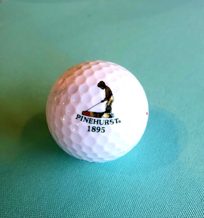 A golf ball with a textured surface placed on a light blue background. The ball features a logo of a person engaged in a golfing stance, accompanied by the text 'PINEHURST 1895'.