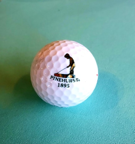 A golf ball with a textured surface placed on a light blue background. The ball features a logo of a person engaged in a golfing stance, accompanied by the text 'PINEHURST 1895'.
