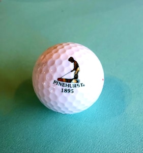 A golf ball with a textured surface placed on a light blue background. The ball features a logo of a person engaged in a golfing stance, accompanied by the text 'PINEHURST 1895'.