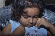 A young child with curly hair is sitting on a intricately designed chair. The child appears to be focused, with eyes cast down, holding a string or cord close to the face.