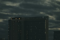 A tall, dark building with illuminated windows stands under an overcast sky. The building is labeled 'Grand Hyatt' and is surrounded by thick, moody clouds.