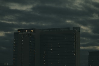 A tall, dark building with illuminated windows stands under an overcast sky. The building is labeled 'Grand Hyatt' and is surrounded by thick, moody clouds.