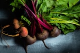 A close-up view of several fresh beets with vibrant red roots and lush green leaves arranged on a textured surface. The beets are partially lit, creating a contrast between the deep red colors and the green foliage.