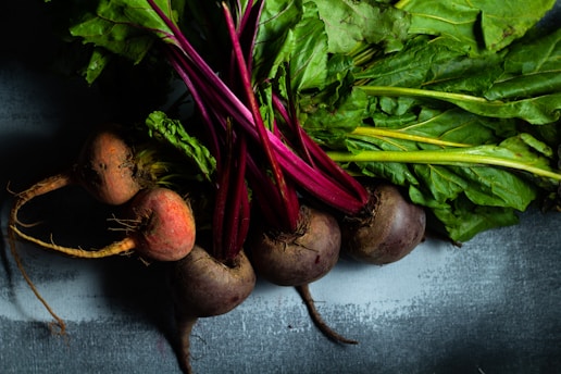 A close-up view of several fresh beets with vibrant red roots and lush green leaves arranged on a textured surface. The beets are partially lit, creating a contrast between the deep red colors and the green foliage.