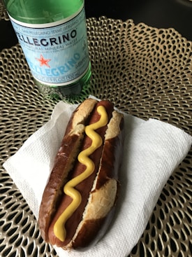 A hot dog with mustard is placed on a white napkin, resting on a decorative woven mat. A bottle of San Pellegrino mineral water is positioned behind it.