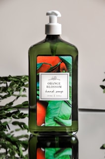 A green bottle of hand soap with a white pump sits on a reflective surface. The label on the bottle features images of oranges and green leaves, indicating the scent is 'Orange Blossom'. Some green foliage is visible in the background, complementing the natural theme of the product.