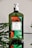 A green bottle of hand soap with a white pump sits on a reflective surface. The label on the bottle features images of oranges and green leaves, indicating the scent is 'Orange Blossom'. Some green foliage is visible in the background, complementing the natural theme of the product.