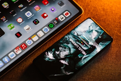 A smartphone and a tablet are placed on a surface with a warm lighting ambiance. The smartphone screen displays an artistic, abstract wallpaper, while the tablet shows a variety of colorful application icons, including Google, Netflix, and others.