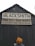A weathered wooden building with a peaked roof features a large sign that reads 'BLACKSMITH' with additional text 'HUB CITY IRON WORKS' and 'BLACKSMITH SHOP-BOILER REPAIR'. A person with reddish hair stands in front of the dark, rustic door.
