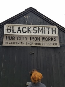 A weathered wooden building with a peaked roof features a large sign that reads 'BLACKSMITH' with additional text 'HUB CITY IRON WORKS' and 'BLACKSMITH SHOP-BOILER REPAIR'. A person with reddish hair stands in front of the dark, rustic door.