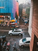 A street scene featuring several vehicles, including white cars and auto rickshaws. The road is wet from the rain, and raindrops are visible falling. A colorful multistory building with shades of blue, red, and pink is on one side, while trees and smaller shops are visible further down the street.