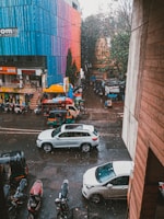 A street scene featuring several vehicles, including white cars and auto rickshaws. The road is wet from the rain, and raindrops are visible falling. A colorful multistory building with shades of blue, red, and pink is on one side, while trees and smaller shops are visible further down the street.