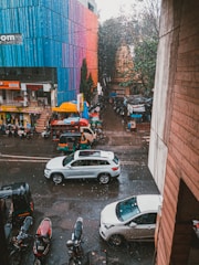 A street scene featuring several vehicles, including white cars and auto rickshaws. The road is wet from the rain, and raindrops are visible falling. A colorful multistory building with shades of blue, red, and pink is on one side, while trees and smaller shops are visible further down the street.