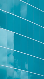 The image features a modern glass building facade with large, reflective blue-tinted glass panels. The panels are arranged in a stylish diagonal alignment, and some clouds are visible reflecting on their surfaces, giving a sense of both structure and openness.