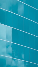The image features a modern glass building facade with large, reflective blue-tinted glass panels. The panels are arranged in a stylish diagonal alignment, and some clouds are visible reflecting on their surfaces, giving a sense of both structure and openness.