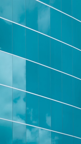 The image features a modern glass building facade with large, reflective blue-tinted glass panels. The panels are arranged in a stylish diagonal alignment, and some clouds are visible reflecting on their surfaces, giving a sense of both structure and openness.