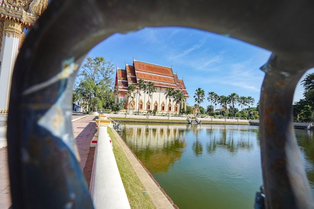 A traditional Thai temple with a distinct red and gold roof is seen across a calm pond. Tall palm trees line the path leading to the temple. The image is framed by a decorative iron structure.