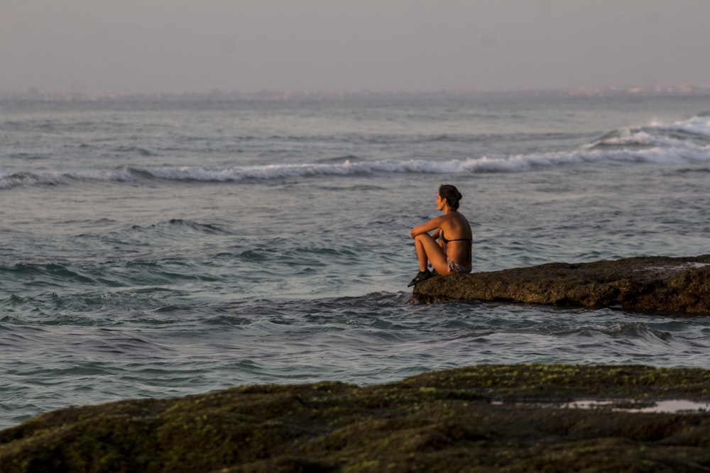 donna in bikini nero che si siede sulla roccia nel mare durante il giorno