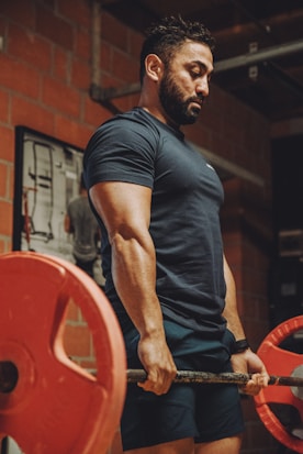 A muscular man is lifting a barbell with large red weights in a gym setting. He is wearing a dark blue shirt and shorts. The background features brick walls and gym equipment, creating an industrial workout atmosphere.
