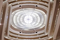 An architectural view of a modern building's interior, featuring multiple circular balconies rising around an open central space. The structure is lit by natural light coming through a large skylight at the top, creating a symmetrical pattern with soft, curved lines and a luxurious atmosphere.