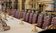 An opulently decorated dining room with a long table lined with plush, high-backed chairs upholstered in rich red leather. The table is adorned with ornate gold candelabras, and the room features intricate gold detailing and dark wood paneling. Classical architectural elements are present, illustrating a sense of luxury and grandeur.