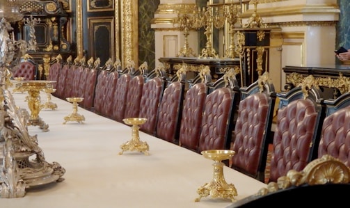 An opulently decorated dining room with a long table lined with plush, high-backed chairs upholstered in rich red leather. The table is adorned with ornate gold candelabras, and the room features intricate gold detailing and dark wood paneling. Classical architectural elements are present, illustrating a sense of luxury and grandeur.
