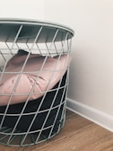 A wire metal basket contains a light pink cushion and a black cushion, situated on a light wooden floor next to a white wall.