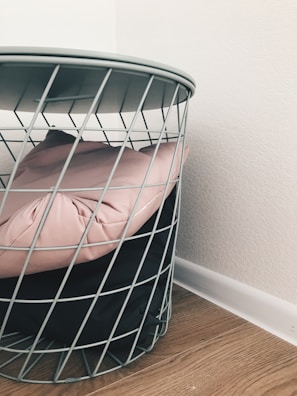A wire metal basket contains a light pink cushion and a black cushion, situated on a light wooden floor next to a white wall.
