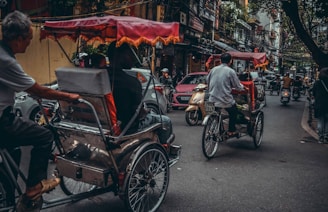 Bicyclists ride rickshaws on a bustling city street surrounded by scooters and cars. The scene is crowded, with intricate power lines hanging overhead and various shops lining the street. The rickshaws have colorful canopies, adding a vibrant touch to the relatively overcast atmosphere.