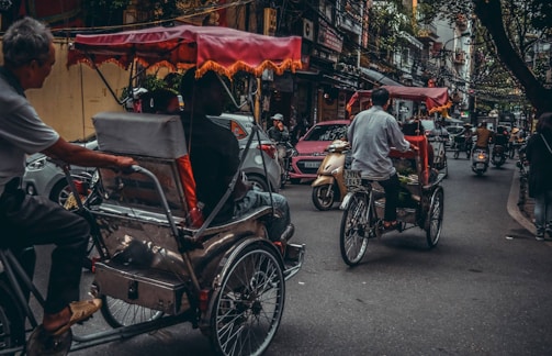 Bicyclists ride rickshaws on a bustling city street surrounded by scooters and cars. The scene is crowded, with intricate power lines hanging overhead and various shops lining the street. The rickshaws have colorful canopies, adding a vibrant touch to the relatively overcast atmosphere.