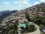 A densely built town with multiple houses crowded closely together on a hillside. The houses are a mix of colors with some having green roofs. The town is surrounded by lush green trees and a winding road can be seen cutting through the buildings. The sky above is clear with a few scattered clouds, and a distant communication tower is visible on a hill.