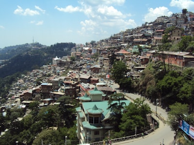 A densely built town with multiple houses crowded closely together on a hillside. The houses are a mix of colors with some having green roofs. The town is surrounded by lush green trees and a winding road can be seen cutting through the buildings. The sky above is clear with a few scattered clouds, and a distant communication tower is visible on a hill.