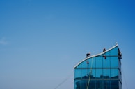 Workers wearing safety gear are positioned on the sloped roof of a modern glass building. The clear blue sky forms the backdrop, emphasizing the architectural lines of the structure. Red safety equipment is visible, indicating a construction or maintenance activity.