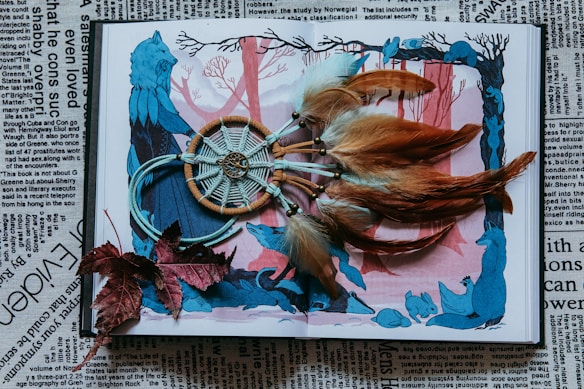 A dreamcatcher with brown and white feathers is placed on an open book featuring an illustration of a blue wolf in a forest scene with red and blue tones. Surrounding the dreamcatcher are a few dried red leaves. The background is filled with newspaper text in black and white.