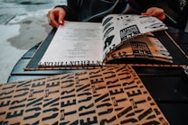 A person with painted nails is sitting at a table, looking at a menu that features bold, graphic text and illustrations. The table is dark and there are other menus or paper items scattered around.