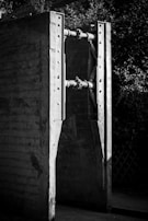 Two large concrete structures connected by metal rods, with bolts securing the connections. The scene is in black and white, highlighting the rugged texture of the concrete and the metallic shine of the rods. The background includes foliage, providing contrast to the industrial elements.