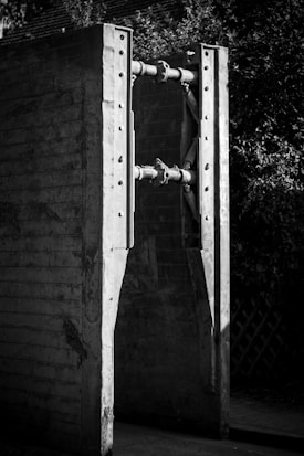 Two large concrete structures connected by metal rods, with bolts securing the connections. The scene is in black and white, highlighting the rugged texture of the concrete and the metallic shine of the rods. The background includes foliage, providing contrast to the industrial elements.