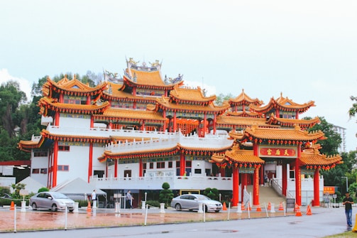 A traditional Chinese temple with vibrant red and gold ornate architecture stands prominently. The multi-tiered roof features intricate detailing and curved eaves, characteristic of Chinese design. Several cars are parked in front, and people walk nearby. Lush greenery surrounds the temple.