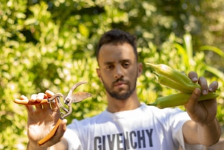 A person is holding a pair of gardening shears in one hand and green peppers in the other while standing outdoors in a lush garden. The background is filled with green foliage, and the person is wearing a white t-shirt.