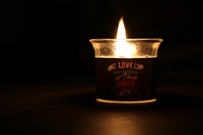 A lit candle in a glass holder casts a warm, glowing light in a dark setting. The candle has an inscription that reads, 'Love one another as I have loved you.' Shadows and reflections can be seen around the candle, emphasizing its gentle illumination.