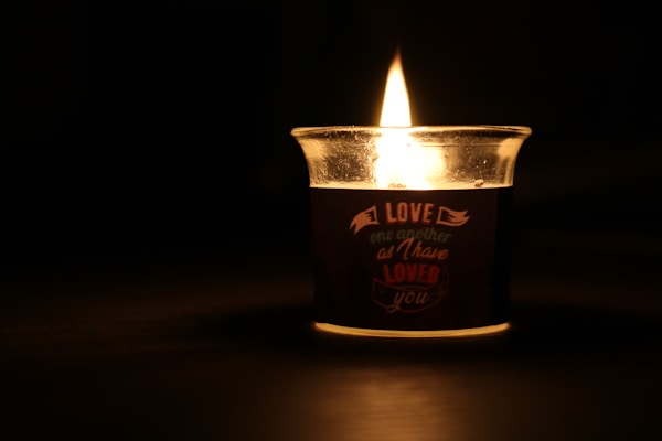 A lit candle in a glass holder casts a warm, glowing light in a dark setting. The candle has an inscription that reads, 'Love one another as I have loved you.' Shadows and reflections can be seen around the candle, emphasizing its gentle illumination.