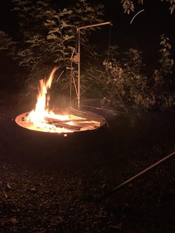 A campfire burning in a fire pit surrounded by pebbles with a metal grilling apparatus nearby. In the background, there are leafy bushes partially illuminated by the fire's glow against a dark night sky.