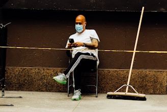 A person wearing a face mask and white shirt sits on a chair against a brown wall, looking at a smartphone. The individual is positioned next to a broom, which is leaning against the wall. A yellow caution tape runs in front of the person.