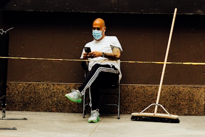 A person wearing a face mask and white shirt sits on a chair against a brown wall, looking at a smartphone. The individual is positioned next to a broom, which is leaning against the wall. A yellow caution tape runs in front of the person.
