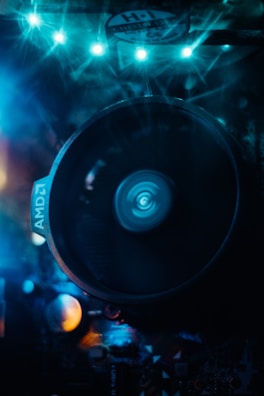 A close-up view of a computer cooling fan with LED lights illuminating the scene in blue. The fan, marked with 'AMD', spins, creating a dynamic blur in the center. Surrounding components are partially visible under the bluish glow, adding a futuristic and high-tech feel to the composition.