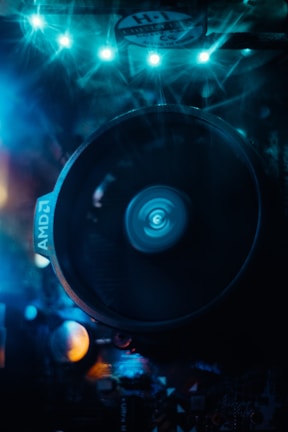 A close-up view of a computer cooling fan with LED lights illuminating the scene in blue. The fan, marked with 'AMD', spins, creating a dynamic blur in the center. Surrounding components are partially visible under the bluish glow, adding a futuristic and high-tech feel to the composition.