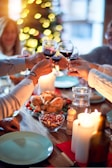 A group of people around a festive dining table clinking glasses of red wine, with a variety of food items including a roasted chicken, mixed nuts, and candles on the table. The background features a decorated Christmas tree with glowing lights.