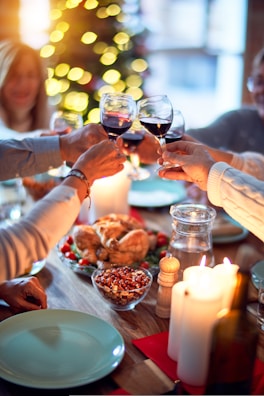 A group of people around a festive dining table clinking glasses of red wine, with a variety of food items including a roasted chicken, mixed nuts, and candles on the table. The background features a decorated Christmas tree with glowing lights.