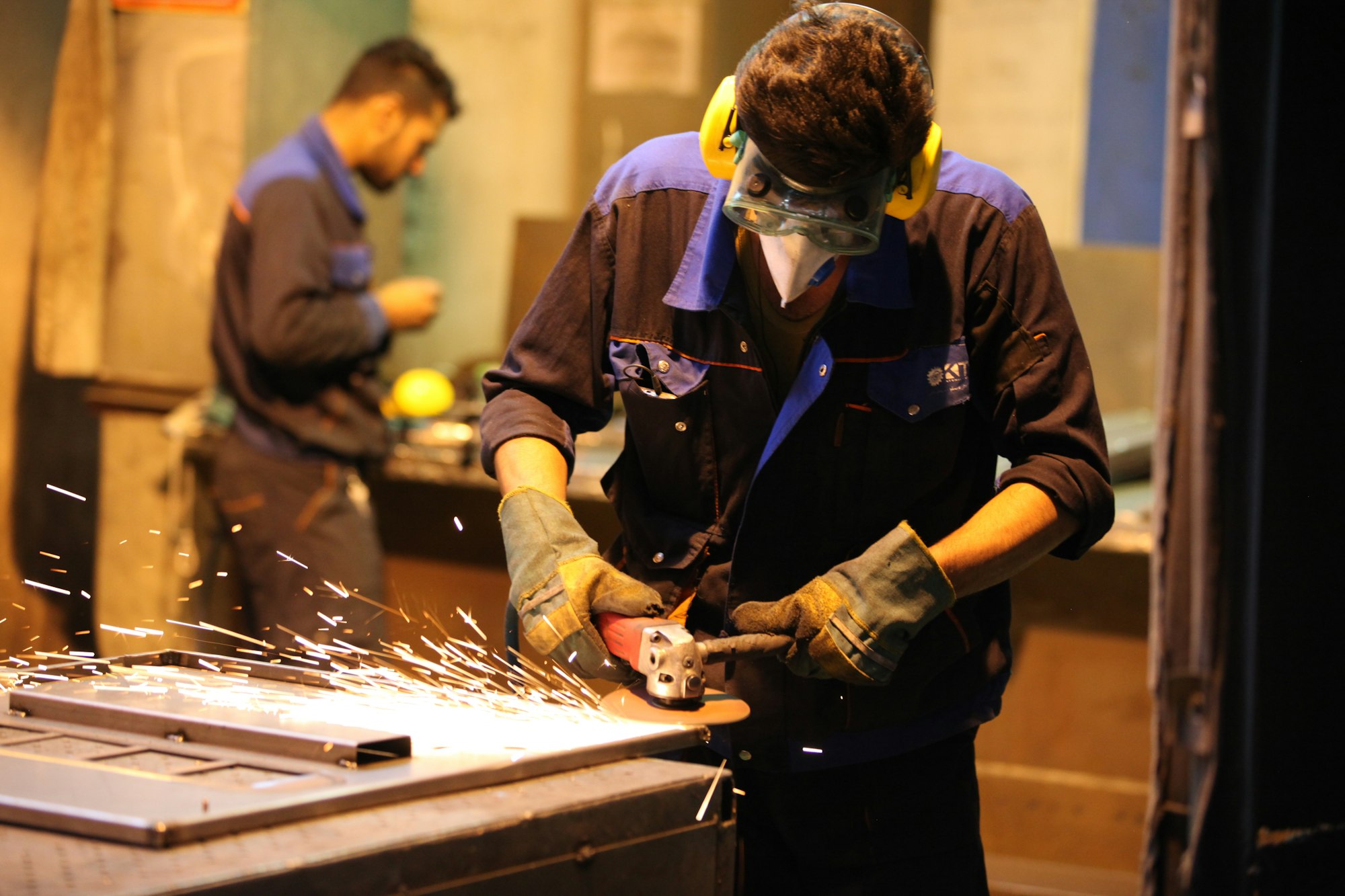 Two workers in safety glasses and ear protection guide an angle grinder across a sheet of metal on a fabrication bench, sparks arcing out toward the floor.