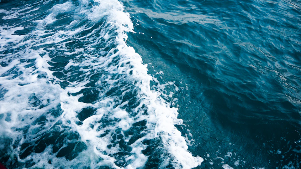 The image depicts a serene scene of a large body of water, possibly an ocean or sea, with its surface disturbed by white foam and waves. The perspective is from above, providing a bird's eye view of the water's surface. The predominant colors in the image are shades of blue and green, reflecting the natural beauty of the scene.